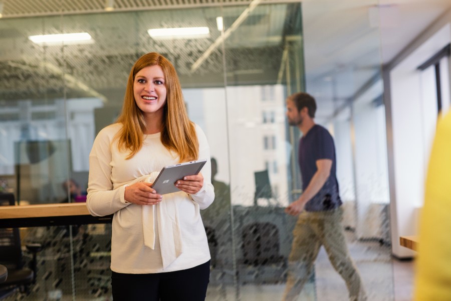 Woman standing, holding a tablet, as a colleague walks by in the background.